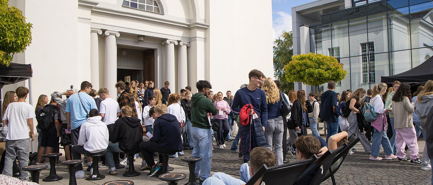Viele Jugendliche sitzen oder stehen auf dem PLatz vor der Kirche beziehungsweise wollen in die Kirche rein. 