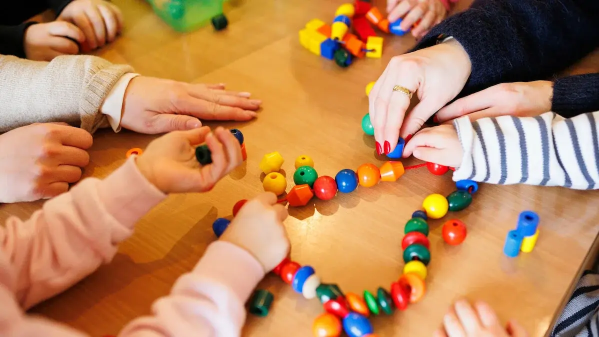 Kita - Kinder beim Spielen. An einem Tisch sitzen einige Kinder, eine Erzieherin und spielen mit einer Kette mit bunten Kugeln und anderen Formen, die auf einer Schnur aufgereiht sind.