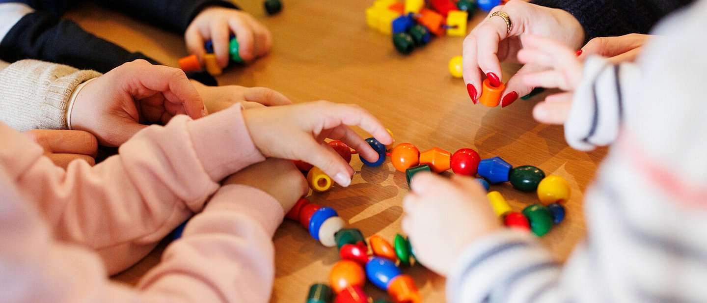 Kita - Kinder beim Spielen. An einem Tisch sitzen einige Kinder, eine Erzieherin und spielen mit einer Kette mit bunten Kugeln und anderen Formen, die auf einer Schnur aufgereiht sind.