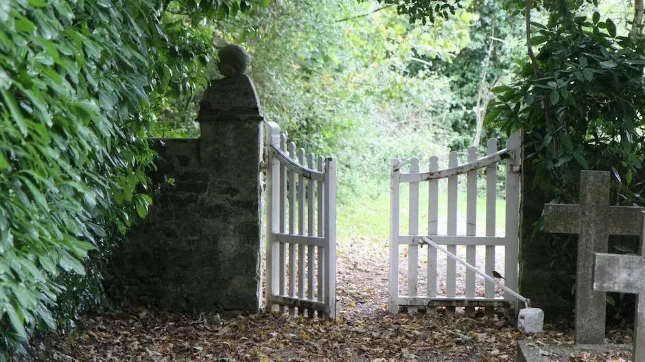 Man erkennt einen Friedhof unter alten Bäumen - rechts ein paar Grabkreuze aus Stein. Ein weißes Holztor ist offen.