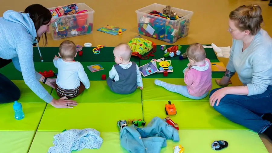 Auf Spielmatten spielen drei Kleinkinder mit Spielsachen. Zwei Mütter sitzen links und rechts.