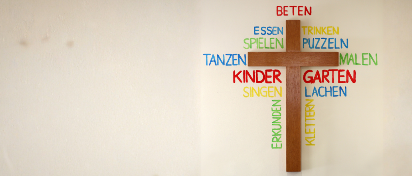 Ein Holzkreuz an einer Wand. Um das Holzkreuz herum stehen Wörter in einer Wortwolke: Kindergarten, Basteln, Spielen, Tanzen und ähnliche Begriffe