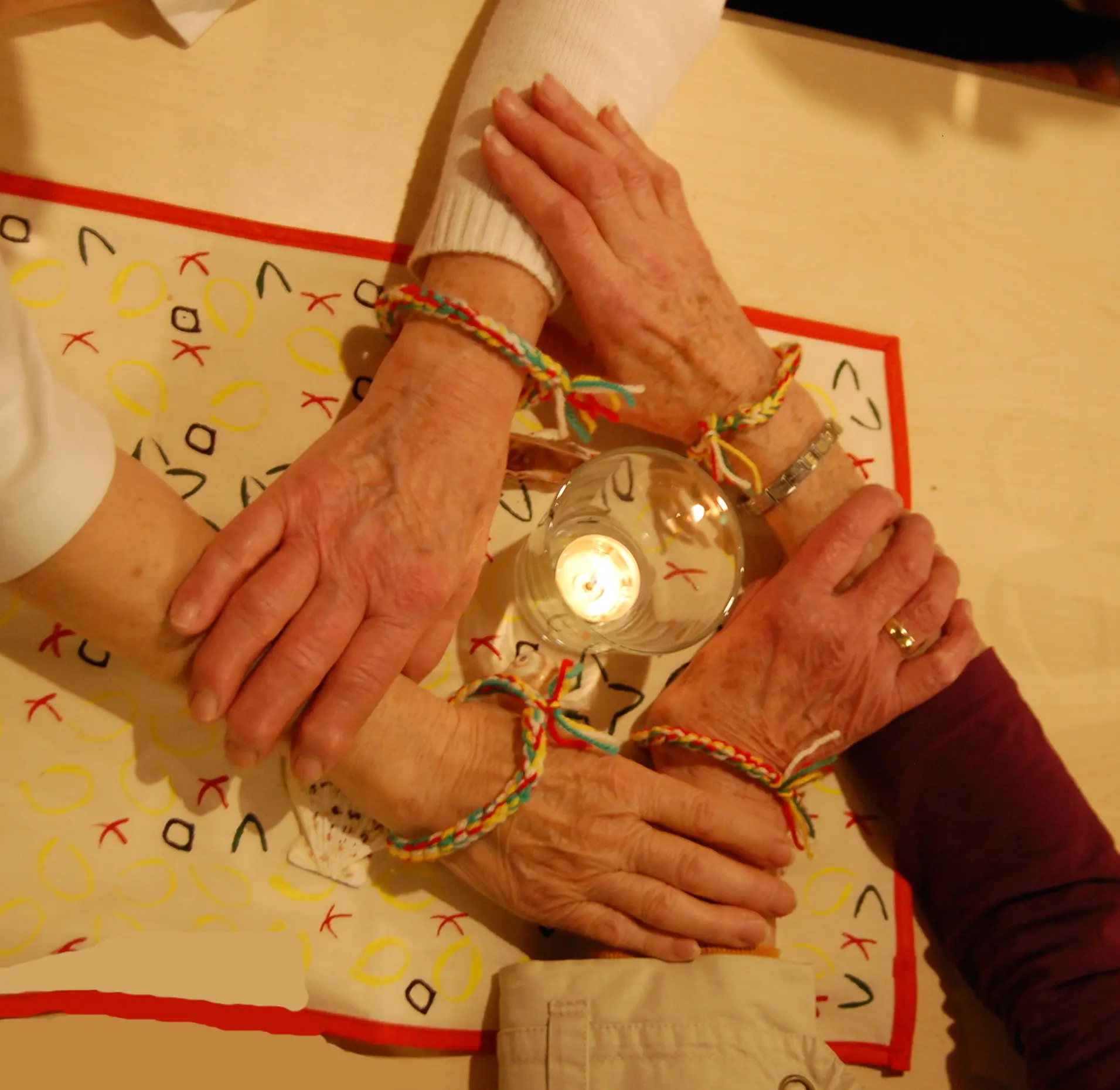 Das Foto zeigt 4 Frauenhände die sich gegenseitig umfassen und um eine brennende Kerze ein Viereck bilden.