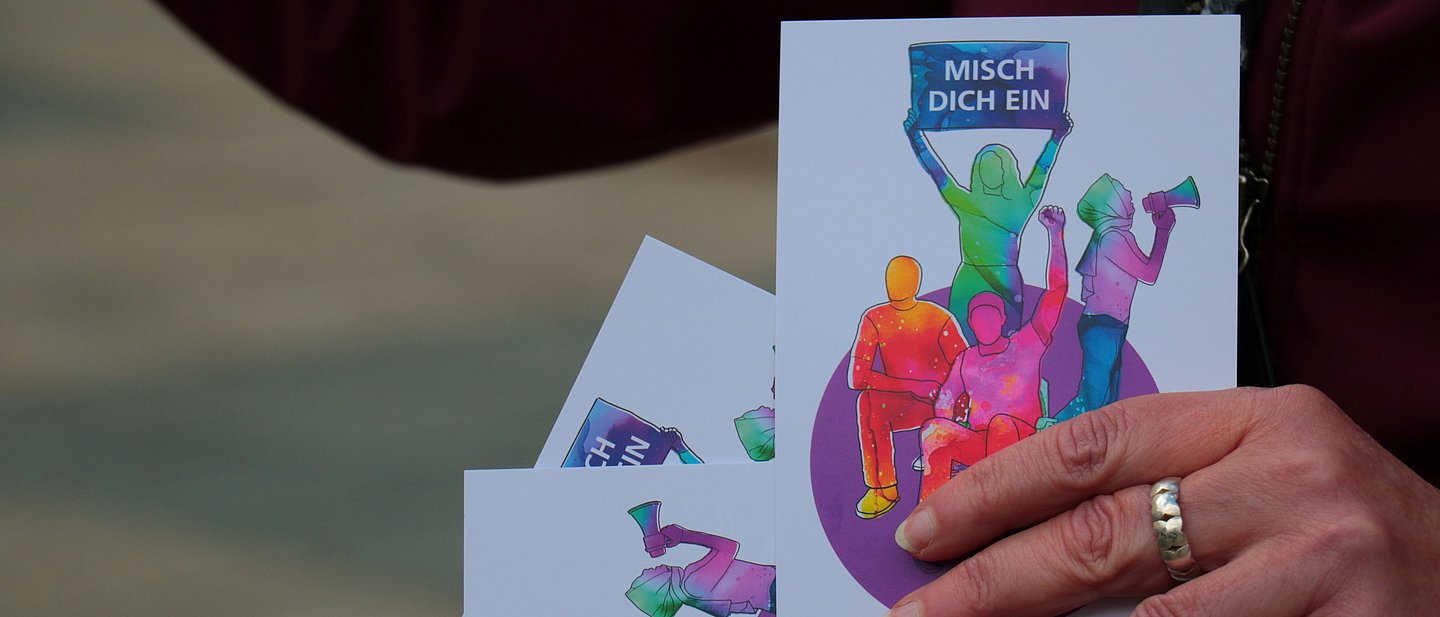 Eine Hand hält Postkarten mit einer bunten Weltkugel und Mensch, die ein Schild "Misch dich ein" hochhalten. 