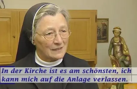 Eine ältere Ordensschwester mit Brille und dunklem Schleier steht in einem Innenraum der Kirche. Im Hintergrund sind ein Holzschrank und eine Heiligenfigur zu sehen. Am unteren Bildrand ist ein eingeblendeter Text zu lesen: „In der Kirche ist es am schönsten, ich kann mich auf die Anlage verlassen.“