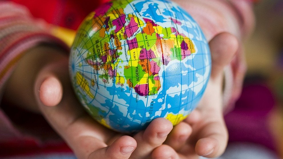 Zwei geöffnete Hände halten einen kleinen Globus mit farbig markierten Ländern. Der Globus zeigt Afrika, Europa und Teile von Asien in kräftigen Farben. Die Hände gehören vermutlich einem Kind, das den Ball behutsam umfasst. Das Bild symbolisiert Fürsorge und weltweite Verbundenheit.