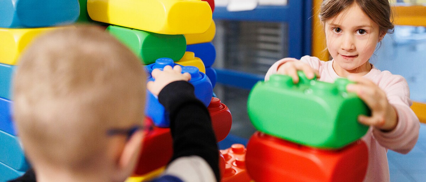 Ev. Kindertagesstätte "Kinder Garten Eden" Gemünden. Ein Junge - von hinten aufgenommen -  baut eine Mauer aus großen, bunten Legosteinen. Das Mädchen baut ebenfalls an der Mauer und schaut zur Kamera.