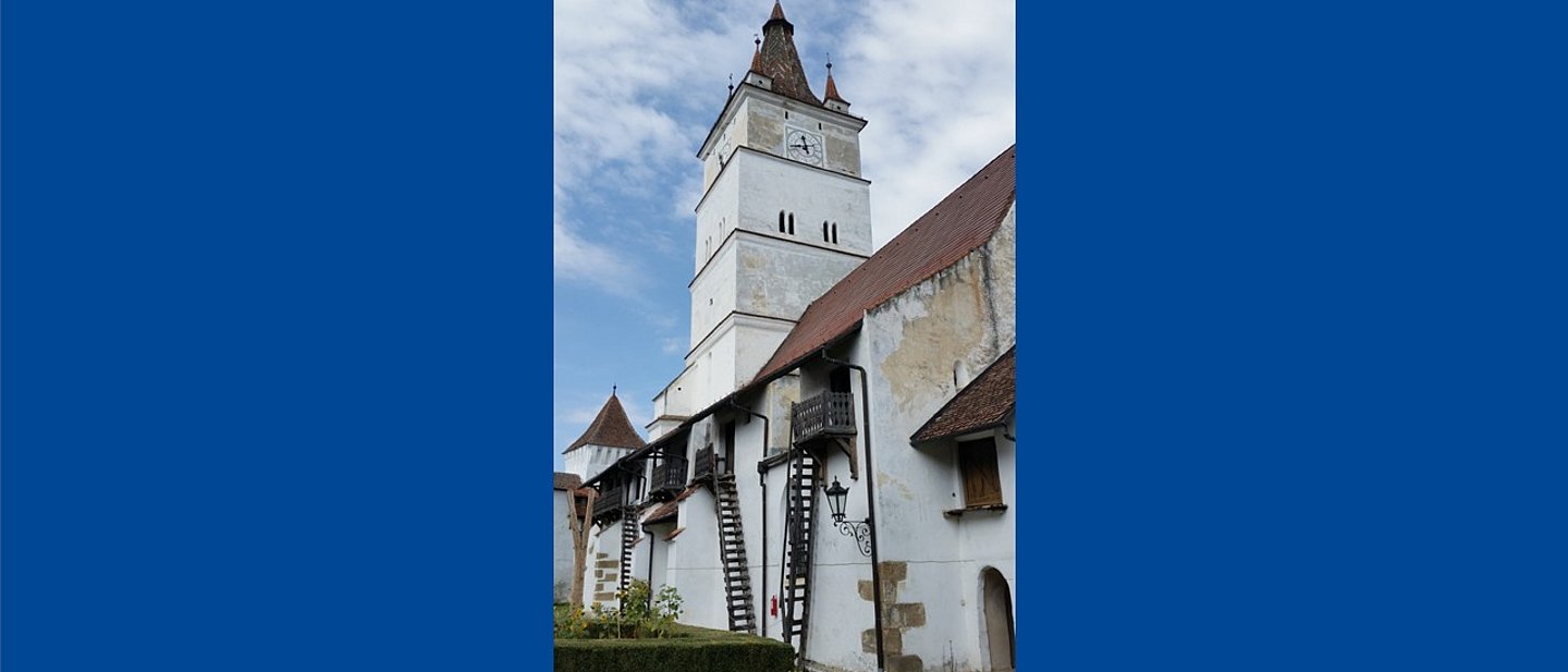 Im Hintergrund ist ein weißer Kirchturm zu sehen. Im Vordergrund ist der Kirchhof und ein Wohngebäude auf dem Kirchengelände.