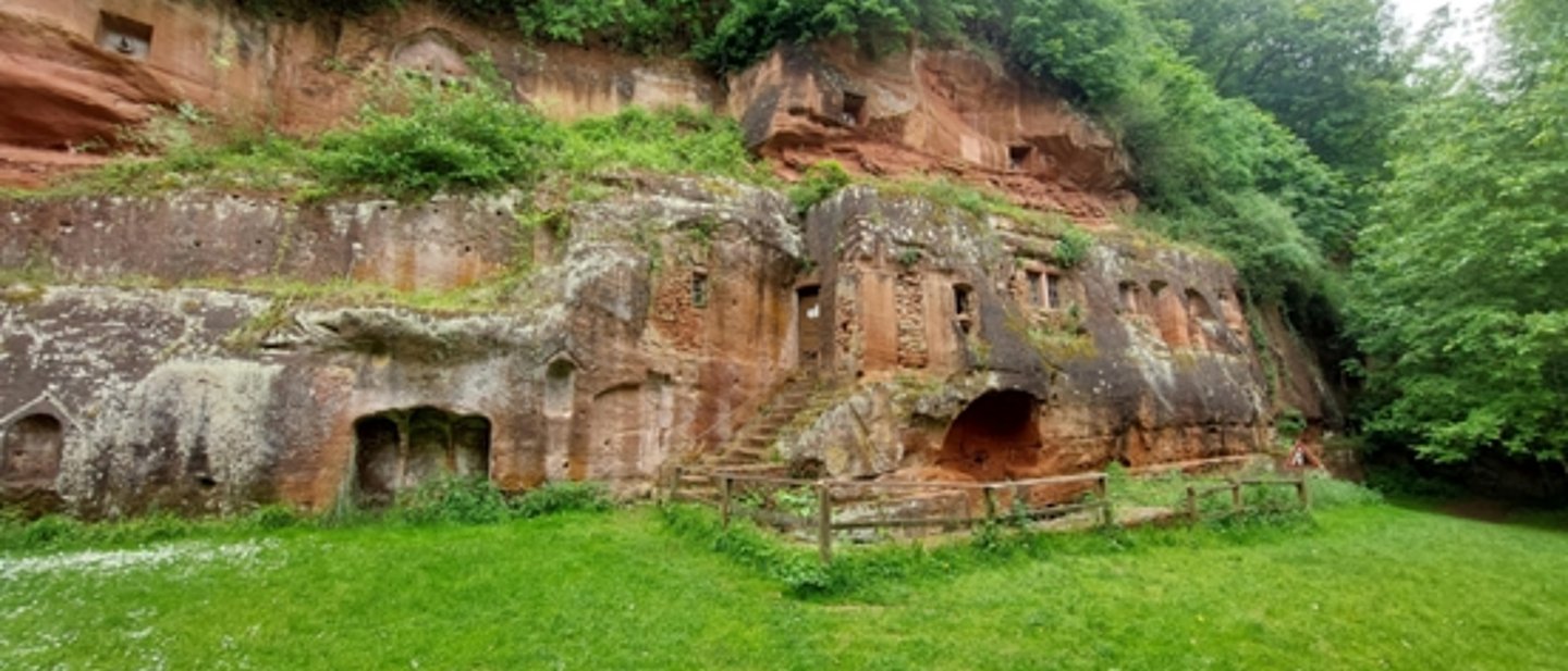 Das Foto zeigt vorne eine grüne Wiese, einen Felsen mit Höhlen, einer Holztür und steinerne Fenster. Darüber wächst eine grüner Wald.