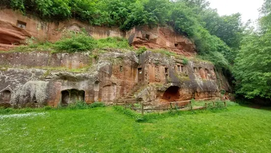 Das Foto zeigt vorne eine grüne Wiese, einen Felsen mit Höhlen, einer Holztür und steinerne Fenster. Darüber wächst eine grüner Wald.