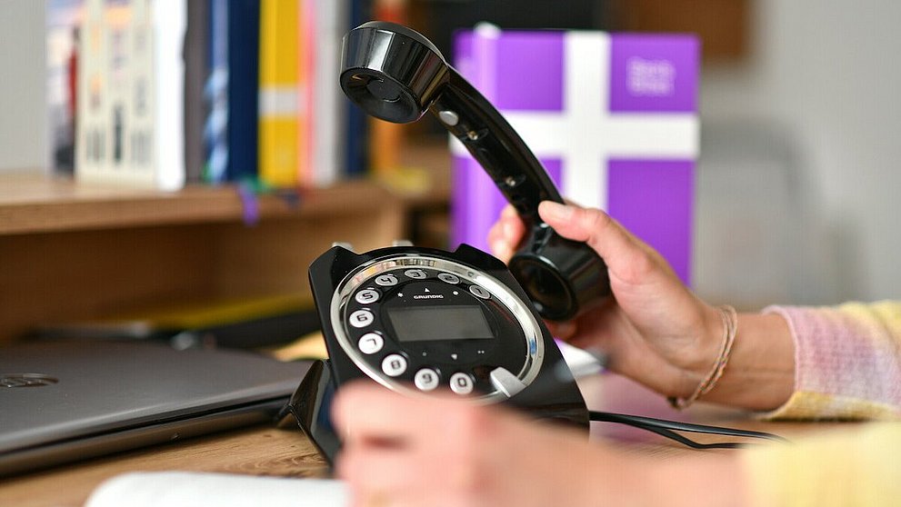 Schreibtisch mit Telefon und einer Hand, die den Telefonhöhrer hält - im Hintergrund in der Unschärfe eine Basisbibel und weitere Bücher.