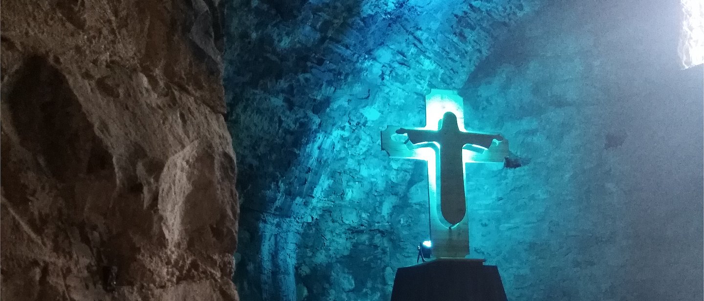 In einem tiefen, dunklen Keller einer Burg leuchtet es blau. Ein Kreuz mit einer Jesusfigur wird angestrahlt. Dahinter ist Licht.