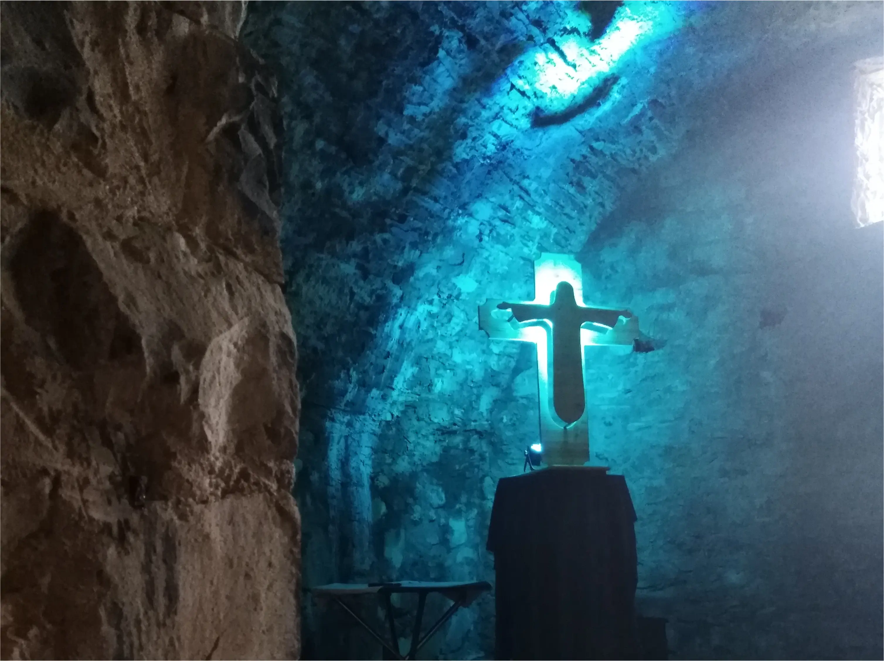 In einem tiefen, dunklen Keller einer Burg leuchtet es blau. Ein Kreuz mit einer Jesusfigur wird angestrahlt. Dahinter ist Licht.