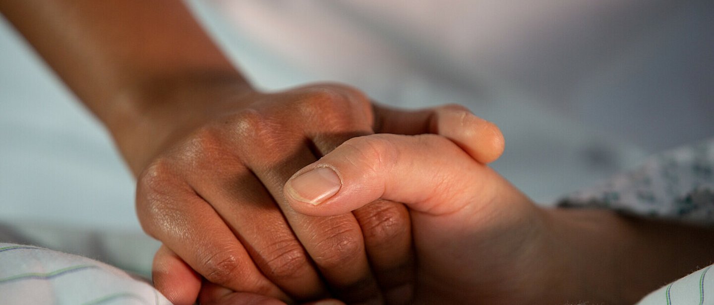 Eine Person liegt auf einem Bett. Ihre Hand wird von einer anderen Person gehalten. Auf dem Bild sind nur die Hände zu sehen.