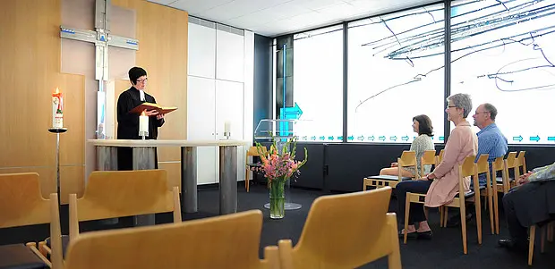 Kleiner Gottesdienstraum mit Holzkreuz an der Wand. Eine Person steht vorne am Altar und liest aus einem Buch vor, während mehrere Besucherinnen und Besucher auf Stühlen sitzen und zuhören. Vorne steht ein Blumenstrauß, große Fenster lassen Tageslicht in den Raum.