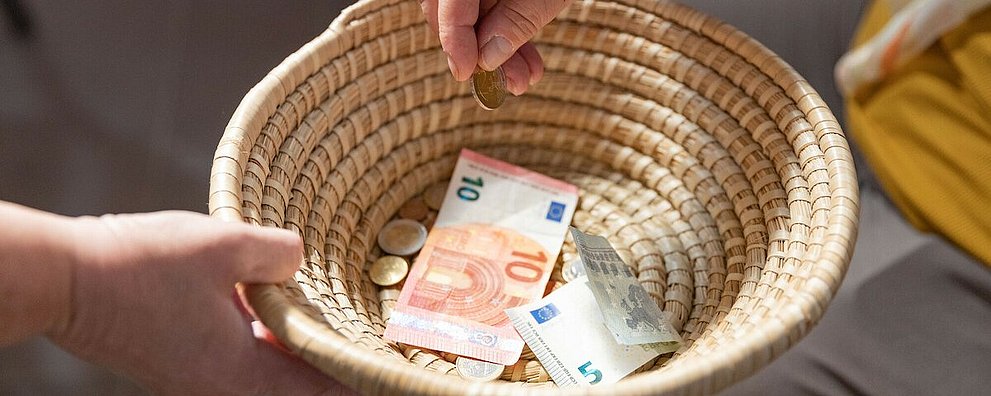Ein geflochtenes Körbchen wird herumgereicht, um Geld einzusammeln. Ein paar Scheine und Münzen befinden sich bereits darin.