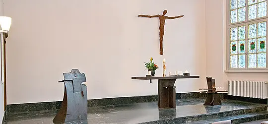 Schlichter Kirchenraum mit Altar und Kruzifix an der Wand; links steht ein steinerner Taufstein, rechts ein Fenster mit Buntglas.