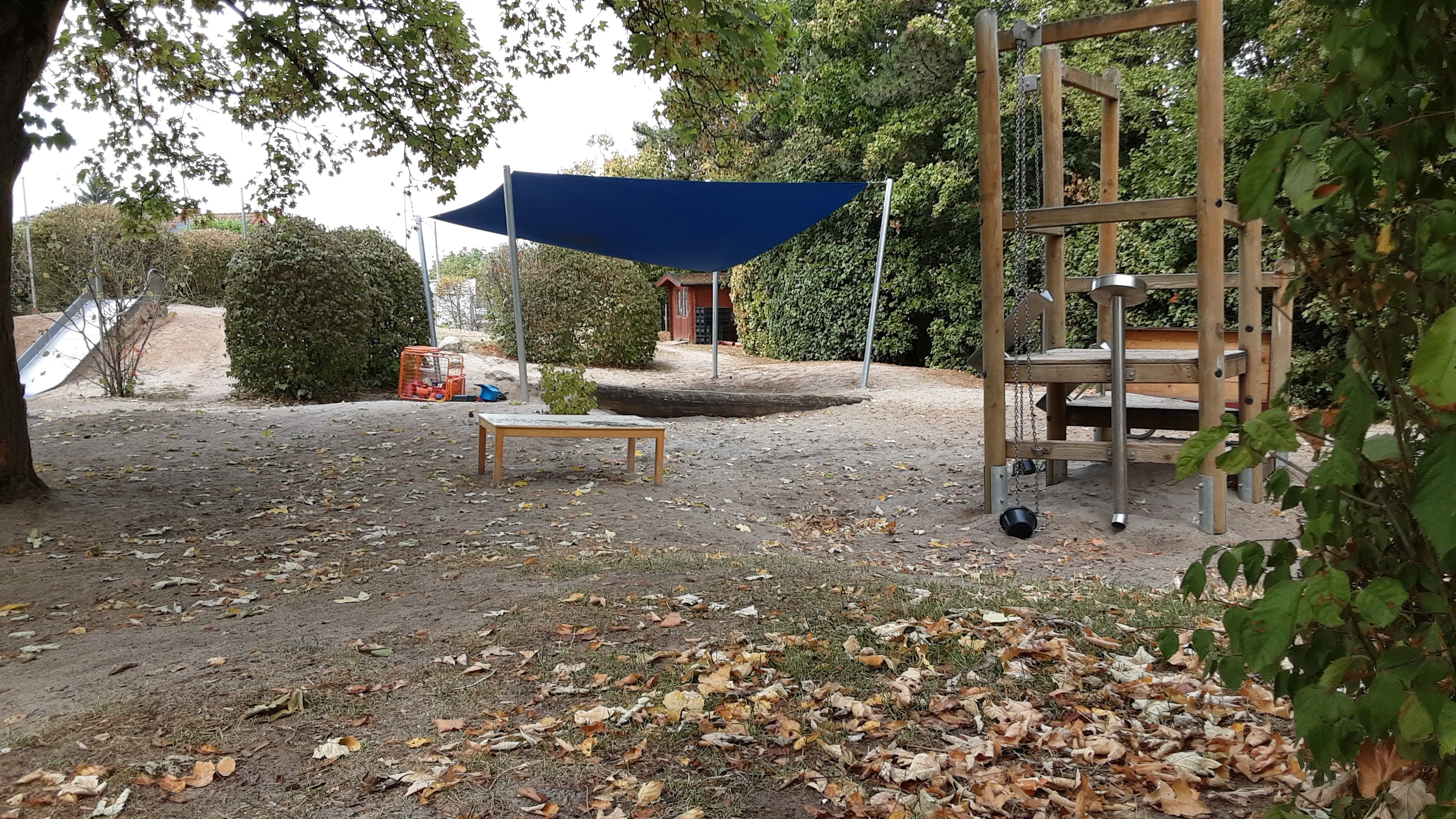 Bild von unserem Außenbereich, in der Mitte des Bildes ein Sankasten mit blauem Sonnensegel, rechts ein Kletterturm, links eine breite Rutschbahn.