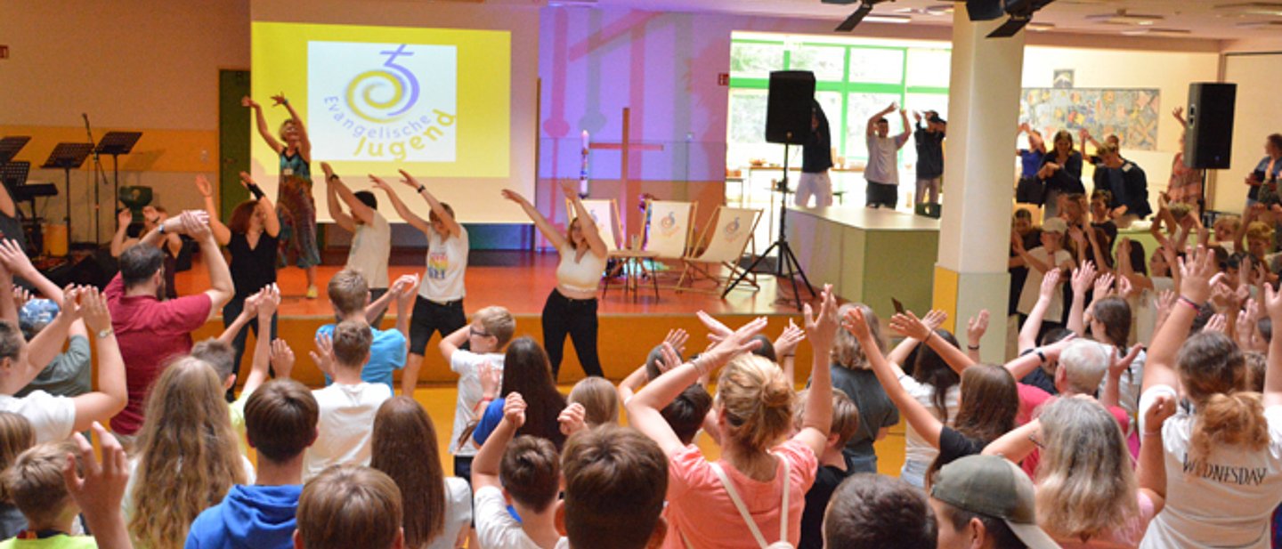 Warm-Up beim Konfitag. Zu sehen ist die Aula einer Schule. Ganz viele Jugendliche schauen auf die Bühne wo andere Jugendliche einen Tanz zur Musik vorführen und die andrern Jugendlichen dann mitmachen und die Hände nach oben bewegen.