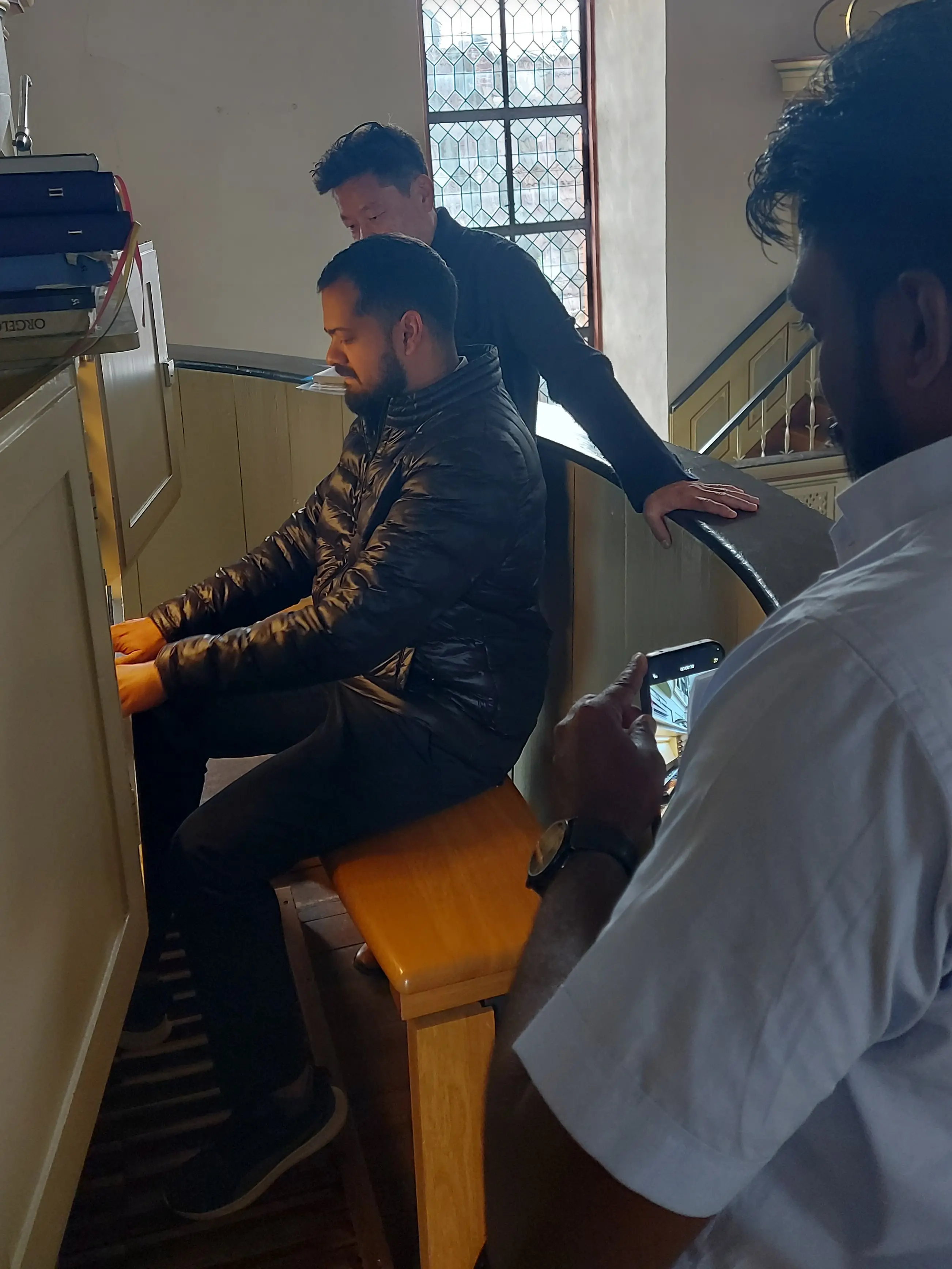 Mann sitzt an einer Kirchenorgel, zwei Männer stehen neben beziehungsweise hinter ihm.
