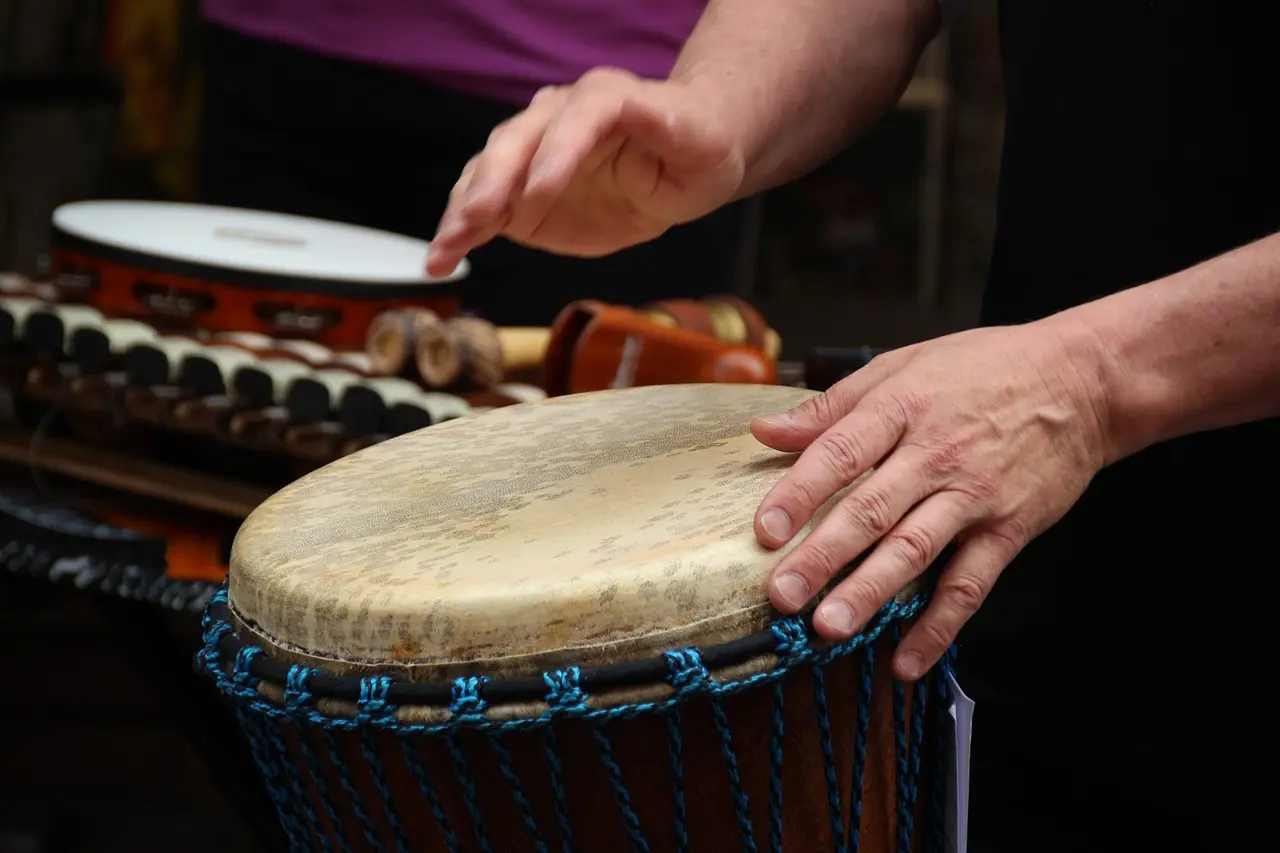 Das Bild zeigt die Hände eines Mannes, die eine Djembe spielen, eine große, kegelförmige Trommel. Im Hintergrund sind weitere Percussion-Instrumente zu sehen.