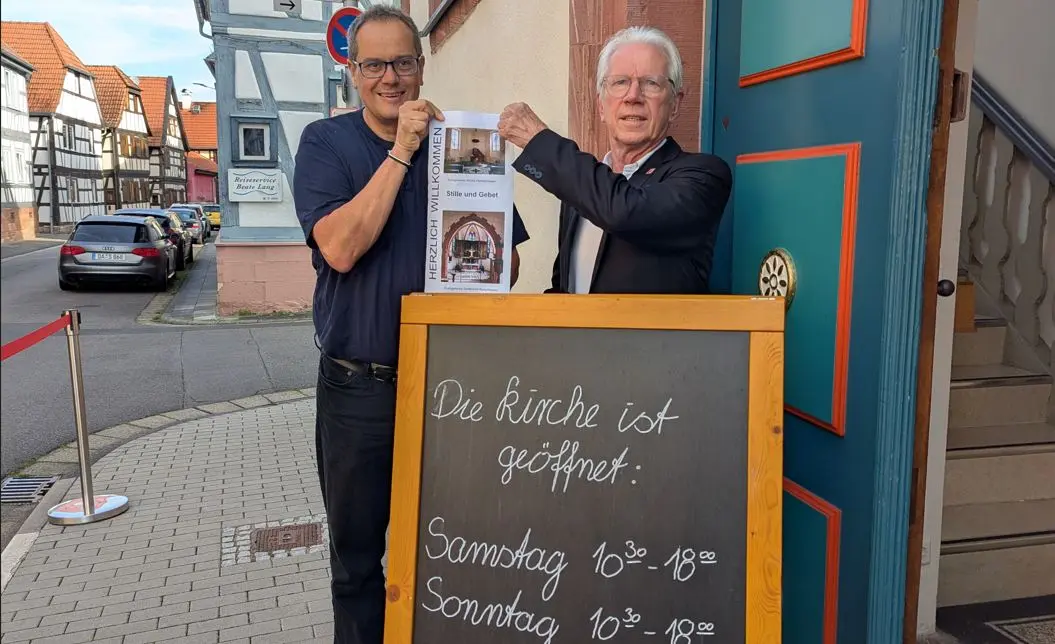 2 Personen stehen vor einem Schild und präsentieren einen Flyer.