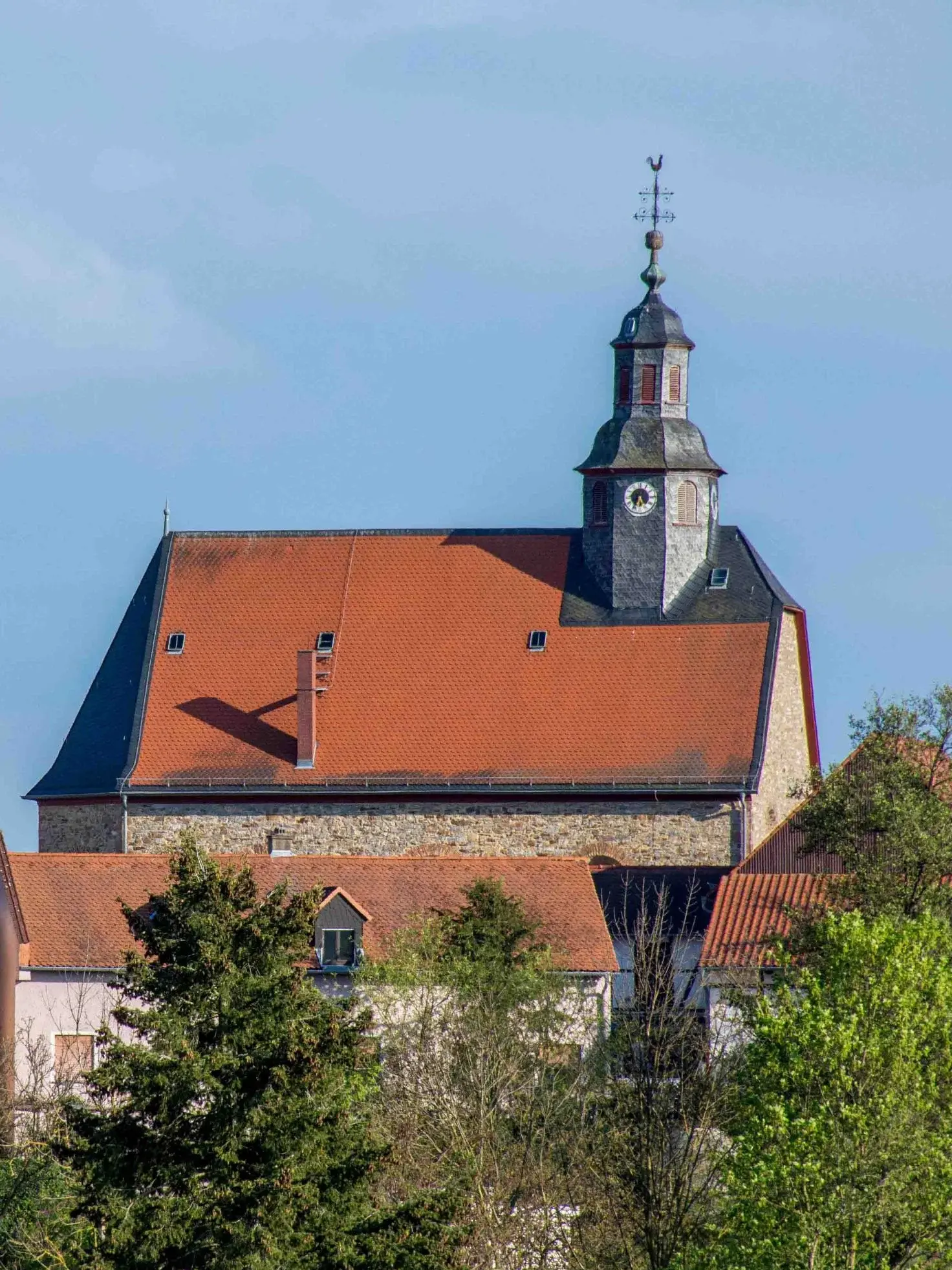 Die Kirche von Burholzhausen an einem sonnigen Tag mit blauem Himmel. Im Vordergrund sind einige Bäume und Büsche zu sehen.