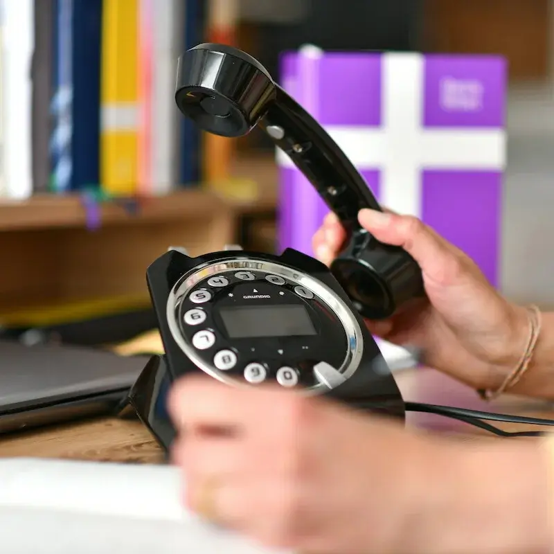 Schreibtisch mit Telefon und einer Hand, die den Telefonhöhrer hält - im Hintergrund in der Unschärfe eine Basisbibel und weitere Bücher.