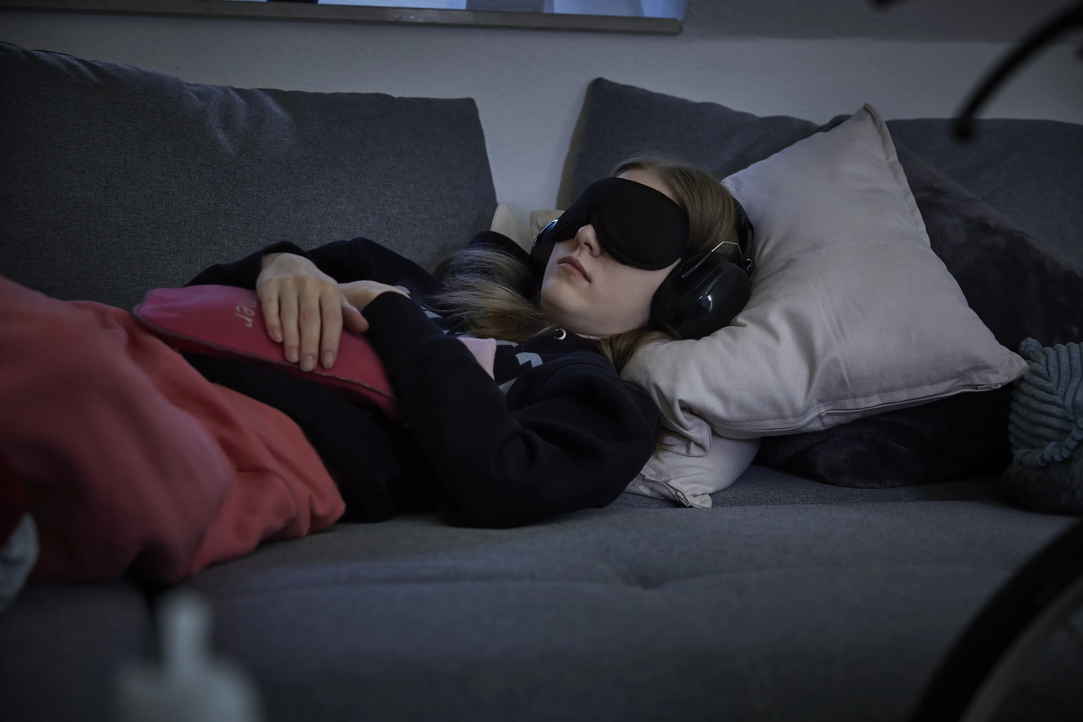 Seite 5: Eine junge Frau liegt auf einem Sofa. In den Händen hält sie eine Wärmflasche mit rotem Bezug. Die Augen sind mit einer schwarzen Schlafbinde bedeckt. Auf den Ohren trägt sie Kopfhörer/Schallschutz.