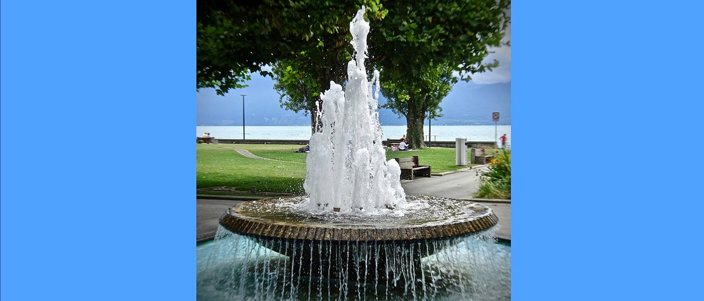 In einem Park steht ein steinerner Brunnen. In der Mitte sprudelt ein hoher Strahl. Das Becken läuft über in einen großen Teich. 