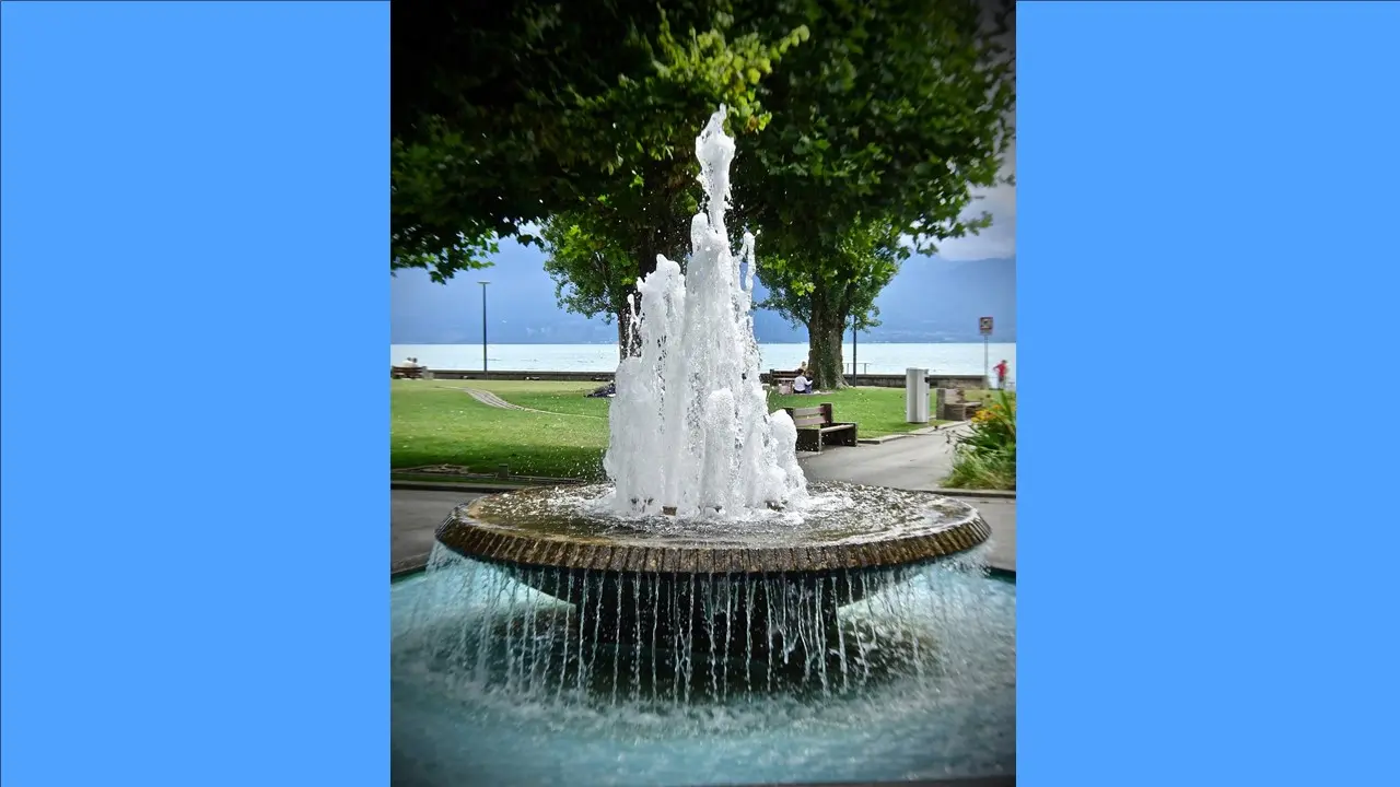 In einem Park steht ein steinerner Brunnen. In der Mitte sprudelt ein hoher Strahl. Das Becken läuft über in einen großen Teich. 