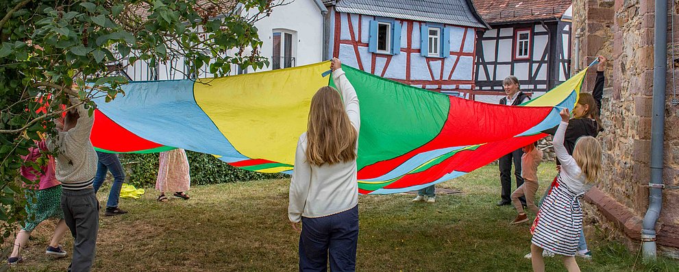 Kinder spielen mit Fallschirm beim Gemeindefest 2025 im Kirchgarten in Burgholzhausen.