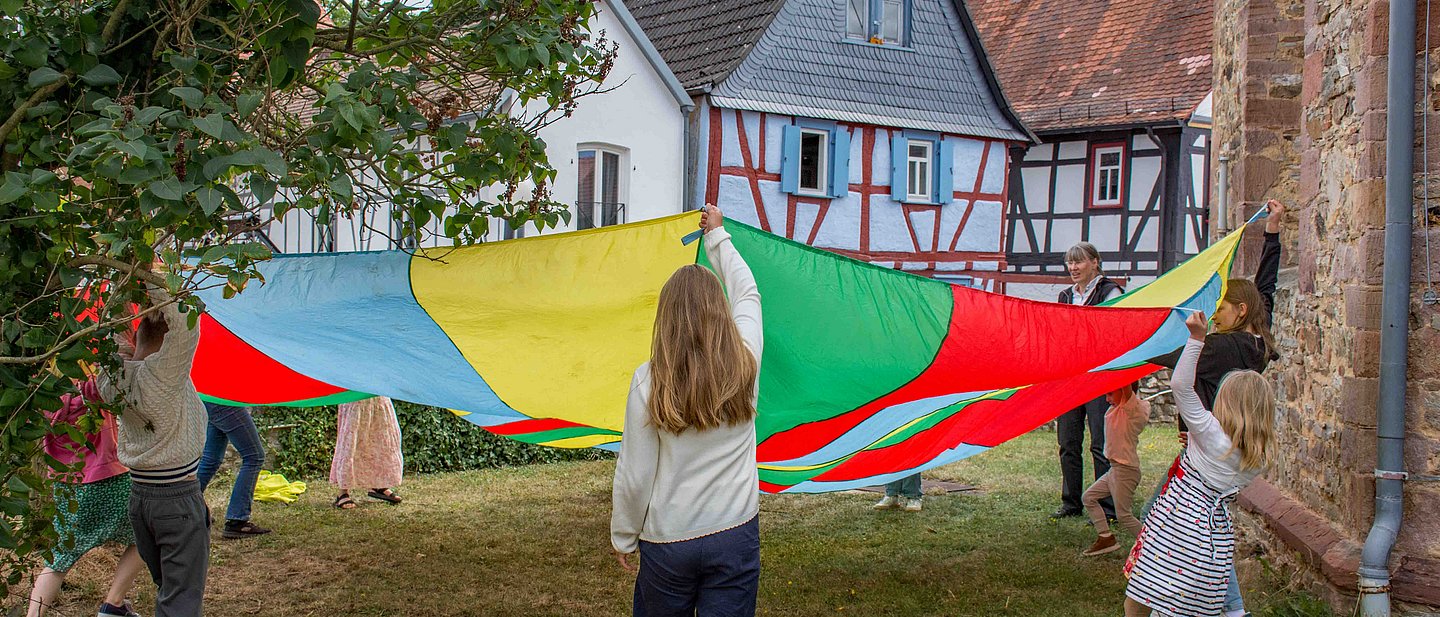 Kinder spielen mit Fallschirm beim Gemeindefest 2025 im Kirchgarten in Burgholzhausen.