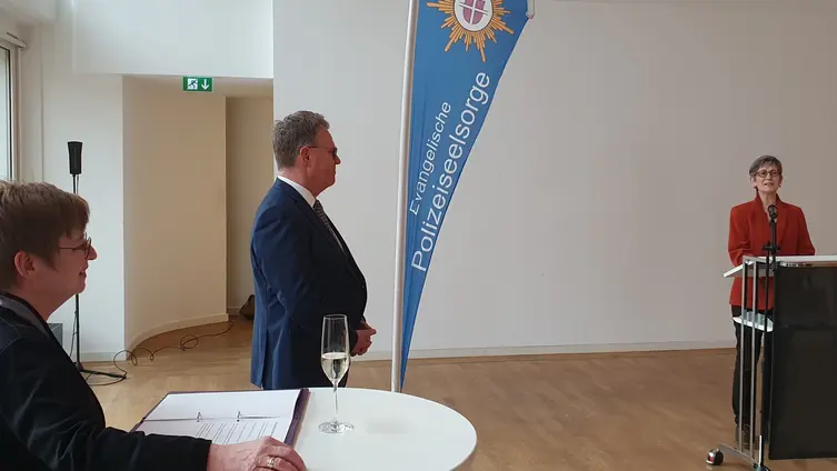 Frau Wirrersteht hinter dem Rednerpult vor einer weißen Wand. Links die beiden Polizeipfarrer Görich-Reinel und Grimm.