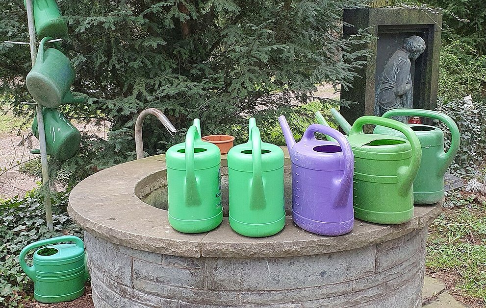 Gießkannen stehen am Rand eines Brunnens. Im Hintergrund ist ein Ständer leerer Gießkannen sowie ein Grabstein mit einer Metallstatue einer Frau zu sehen.