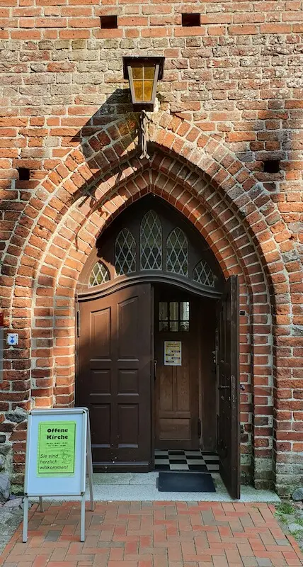 Eingang einer Backsteinkirche mit offener Tür. Über dem Eingang hängt eine Laterne und davor steht ein Schild mit einer Einladung zum Eintritt.