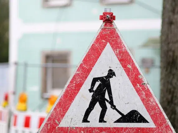 Verkehrsschild Baustelle dreieckig mit rotem Rahmen. Im Hintergrund unscharf das Pfarrhaus