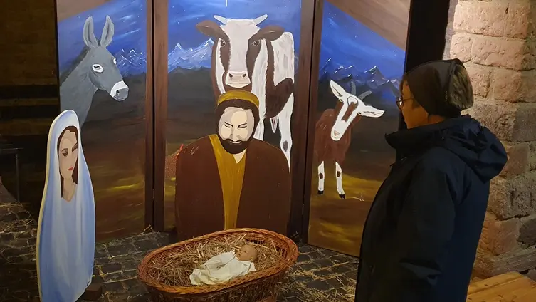 Eine Person betrachtet große Figuren von Maria und Joseph. Die schauen auf einen kleinen Korb aus Bast mit einer Puppe als Jesuskind. Im Hintgrund große Bilder von Ochs und Esel.