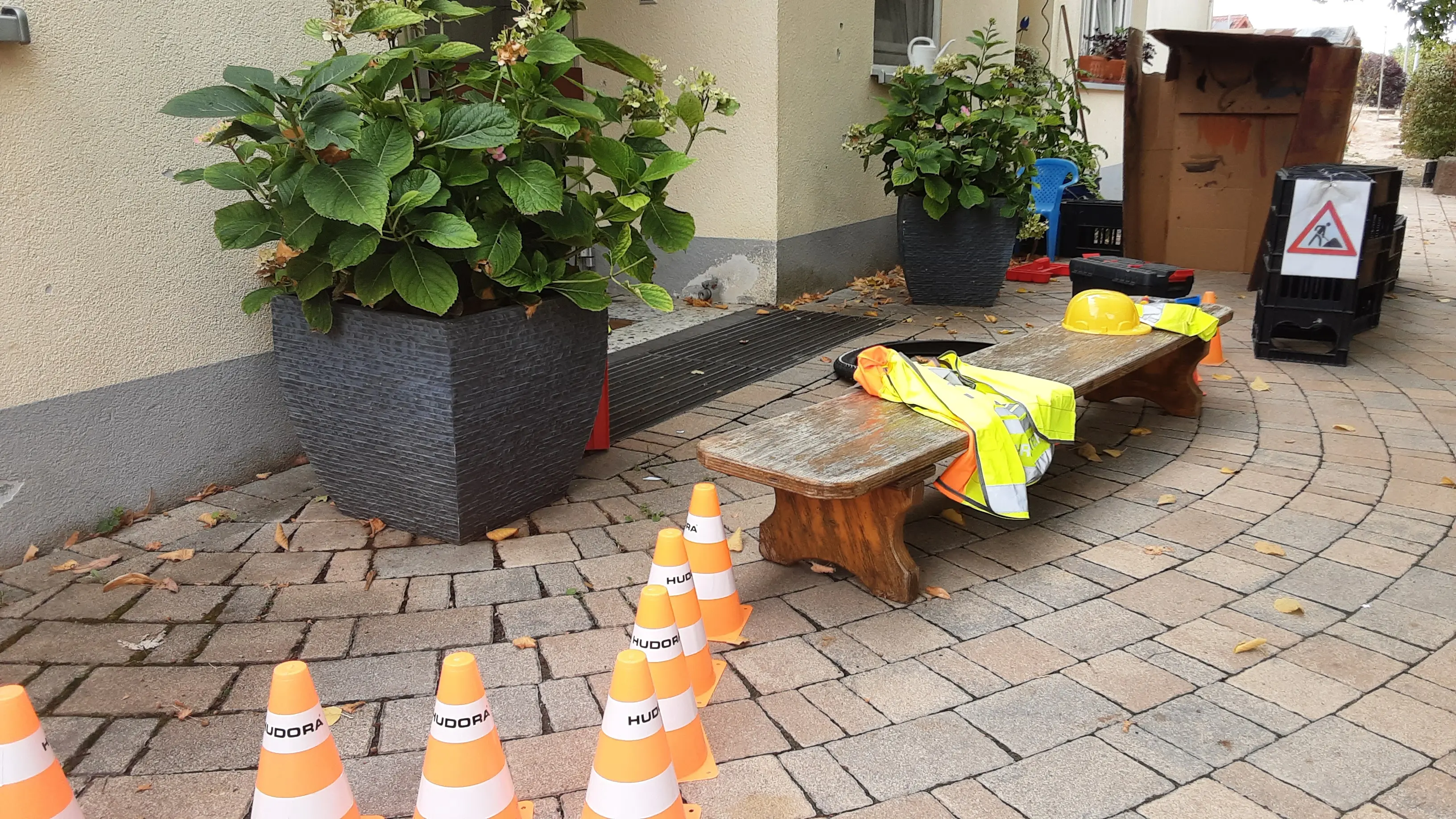 DIe Kinder haben vor dem Haupteingang eine Baustelle mit Pylonen im Vordergrund, einer Bank mit Warnwesten und Helm, Wasserkisten und einem großen Pappkarton aufgebaut.