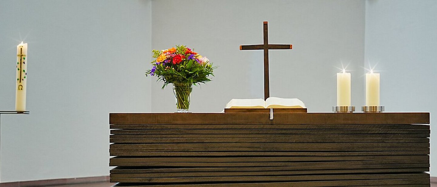 Modern gestalteter Altar auf dem ein Kreuz, eine Altarbibel, Blumenschmuck und zwei Kerzen stehen.
