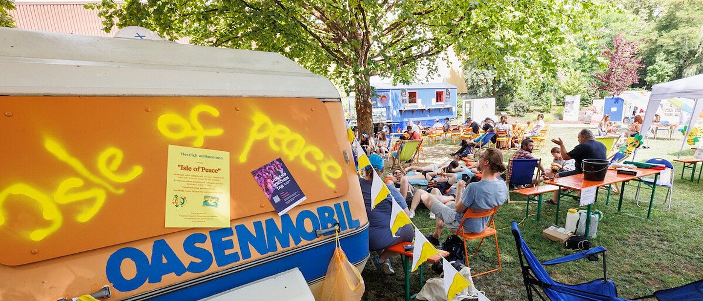 Man sieht im Vordergrund das Oasenmobil und im Hintergrund Jugendliche auf Bänken und Stühlen