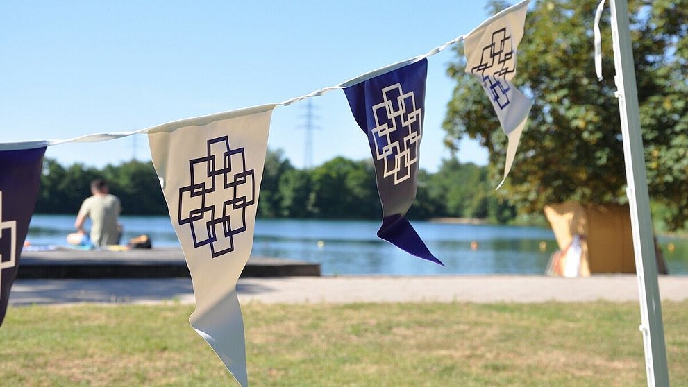 Zu sehen sind Wimpel mit dem Logo der EKHN beim Tauffest in Bensheim. Im Hintergrund befindet sich ein Badesee.