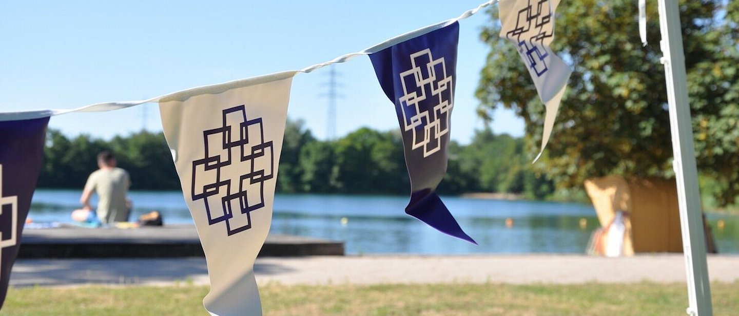 Zu sehen sind Wimpel mit dem Logo der EKHN beim Tauffest in Bensheim. Im Hintergrund befindet sich ein Badesee.