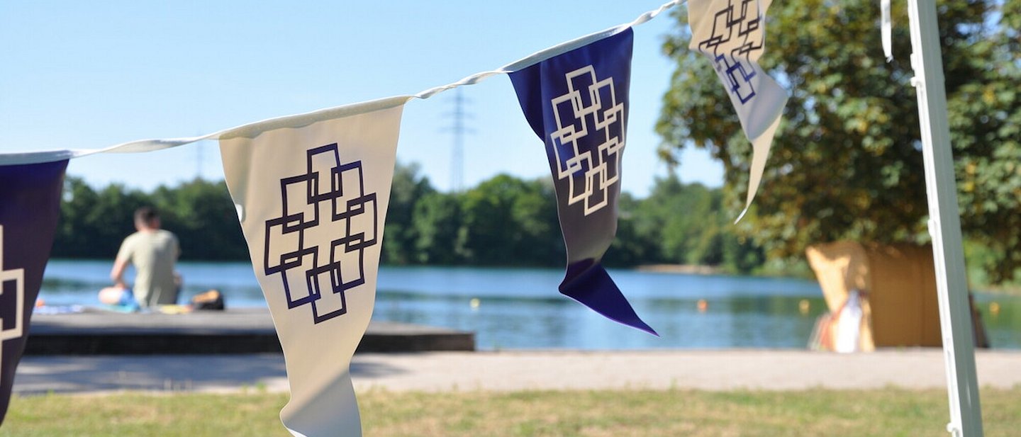Zu sehen sind Wimpel mit dem Logo der EKHN beim Tauffest in Bensheim. Im Hintergrund befindet sich ein Badesee.