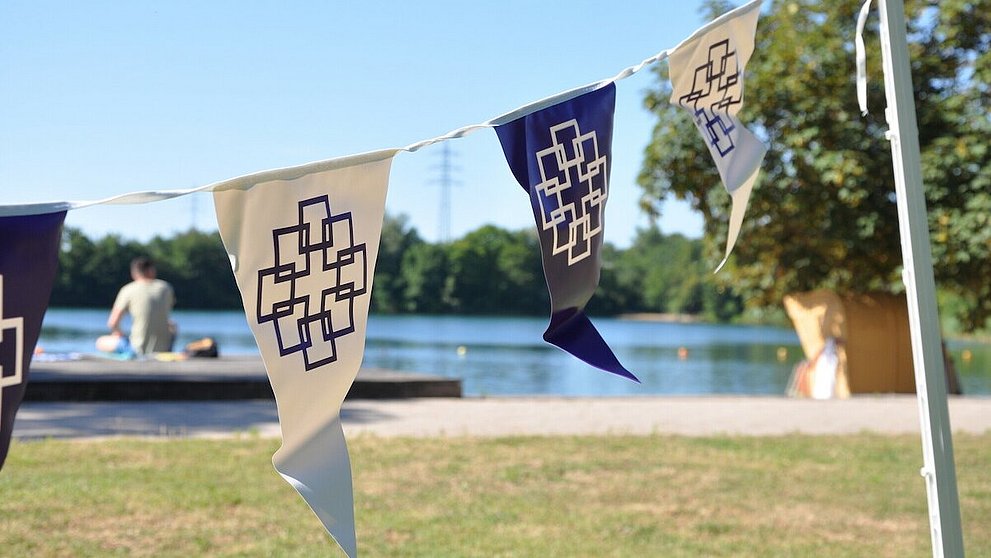Zu sehen sind Wimpel mit dem Logo der EKHN beim Tauffest in Bensheim. Im Hintergrund befindet sich ein Badesee.