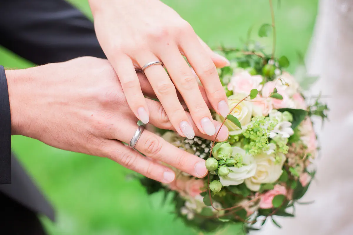 Das Bild zeigt zwei ineinander verschlungene Hände mit Eheringen und Blumenstrauß.