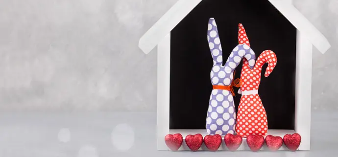 Zwei stoffgenähte Hasenfiguren mit bunten Punkten stehen eng nebeneinander in einem kleinen weißen Häuschen. Vor ihnen liegen mehrere rote Herzdekorationen in einer Reihe. Die Szene wirkt verspielt und liebevoll.