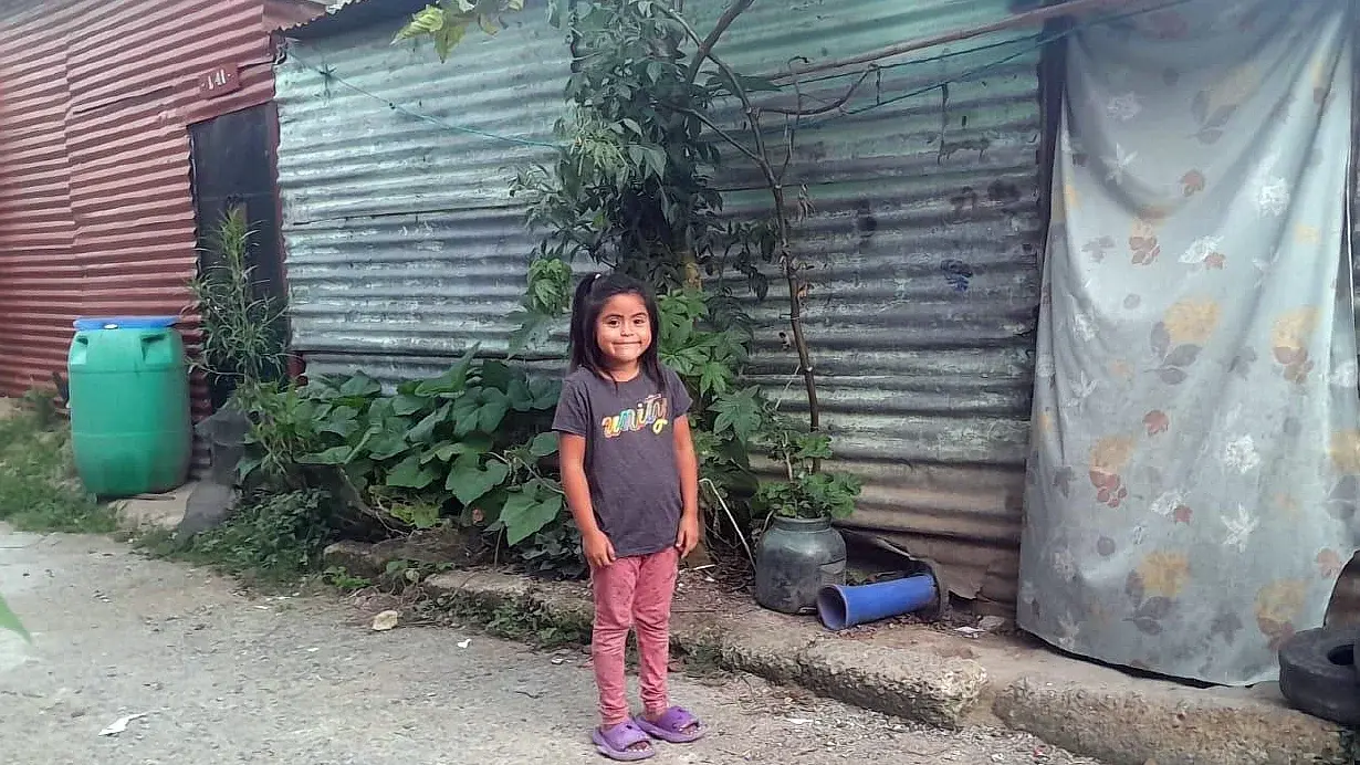 Unser neues Patenkind, die 6jährige Stephany, steht vor ihrem Zuhause, einer Wellblechhütte. Sie lächelt in die Kamera. 