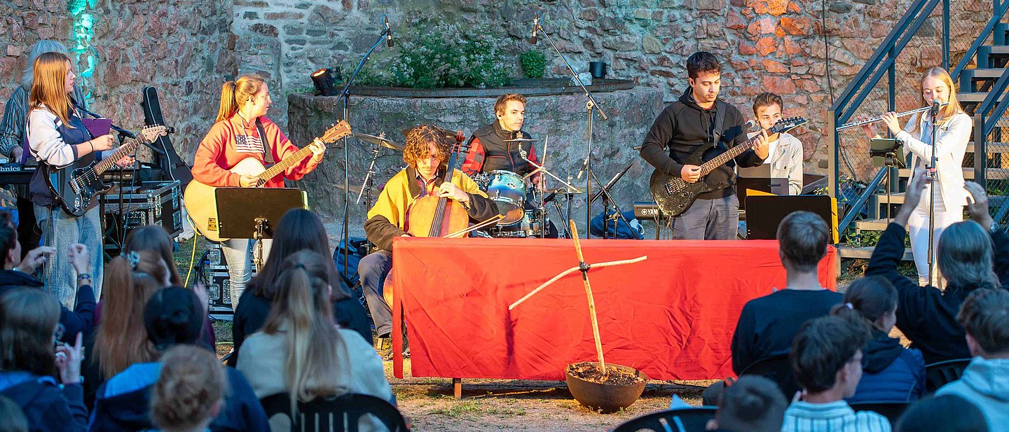 Jugendliche und junge Erwachsene spielen vor Publikum ein Konzert mit Gitarren, Cello, Schlagzeug und Querflöte vor einer beleuchteten Steinmauer.