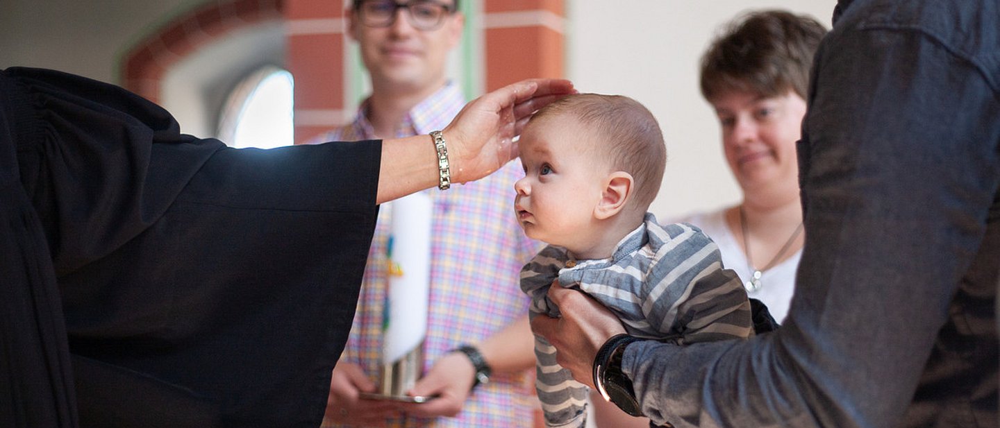 Ein Pfarrer tauft ein Baby, das ein Taufpate über das Taufbecken hält.
